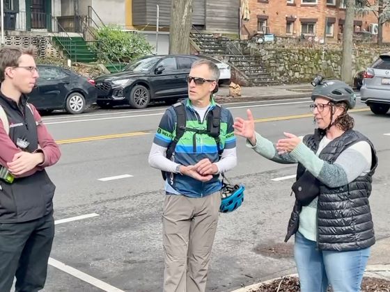 Councilmember Nadeau gestures as she speaks with a man with bicycle helmet and another person at the edge of a road with cars parked across the street.