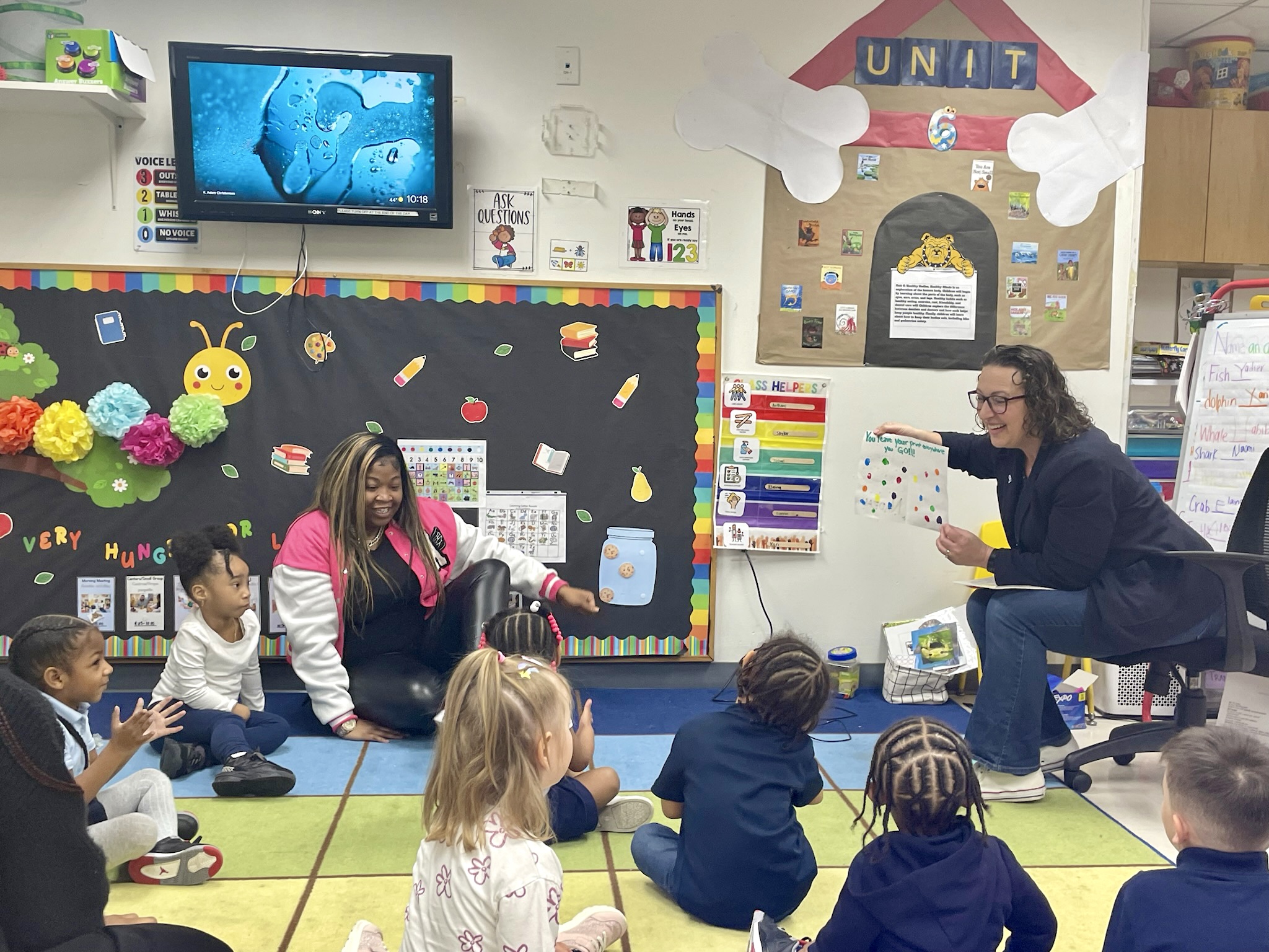 Photo of preschool classroom with students and teacher sitting on a rug against a black bulletin board decorated with a catepillar. Councilmember Brianne K. Nadeau pictured at right holding up a picture big, smiling and reading aloud.