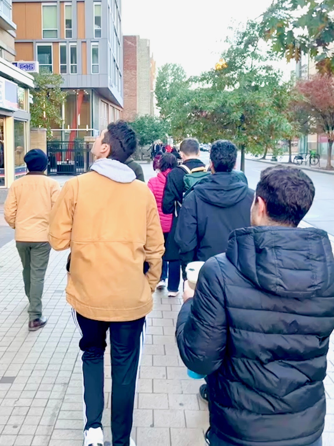 A group of residents, wearing coats on a chilly day, walk down a sidewalk away from the camera. One man is looking left toward a building.