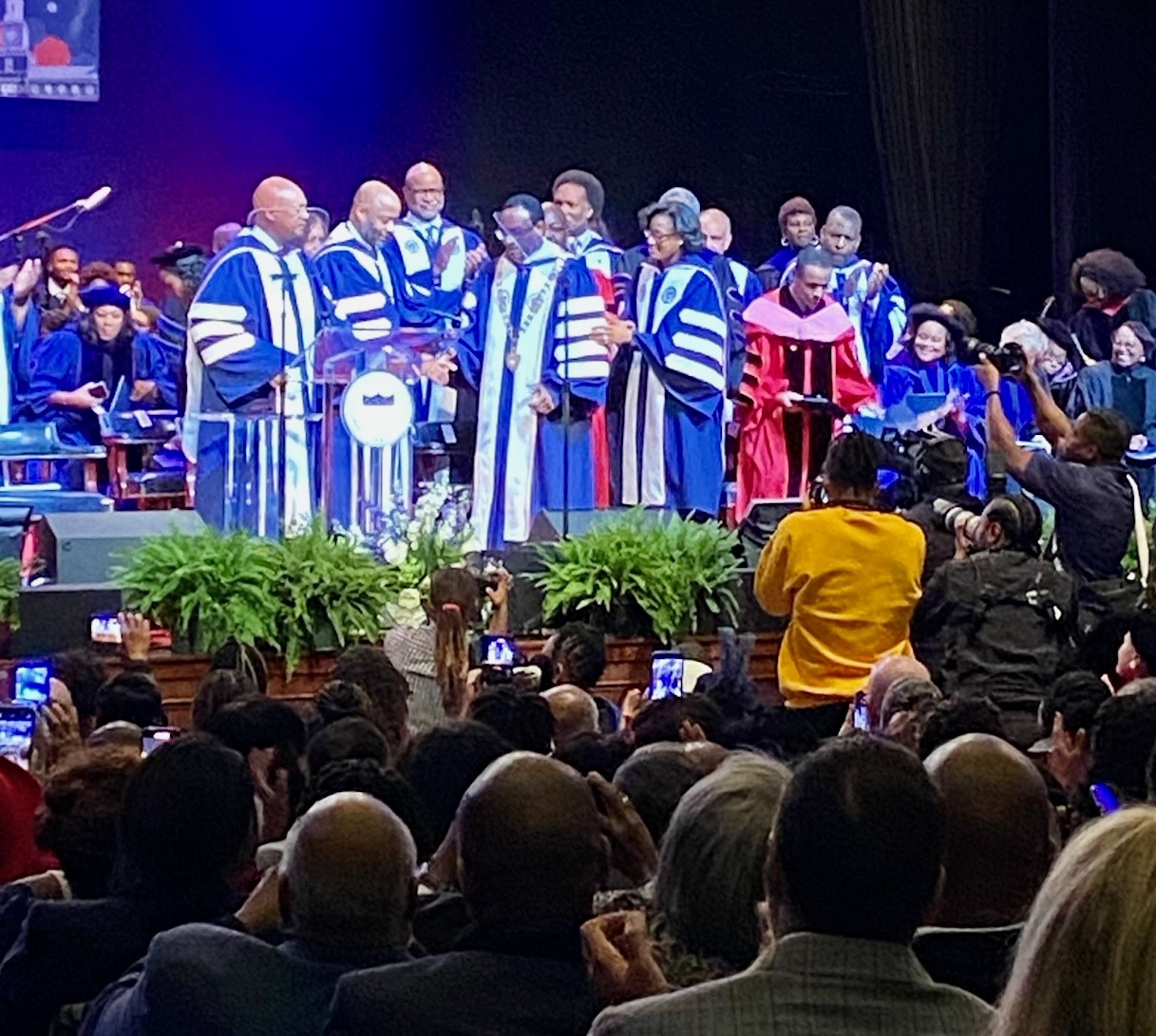President Vinson is on the stage in academic garb, surrouned by members of the faculty. Many audience members are seated.