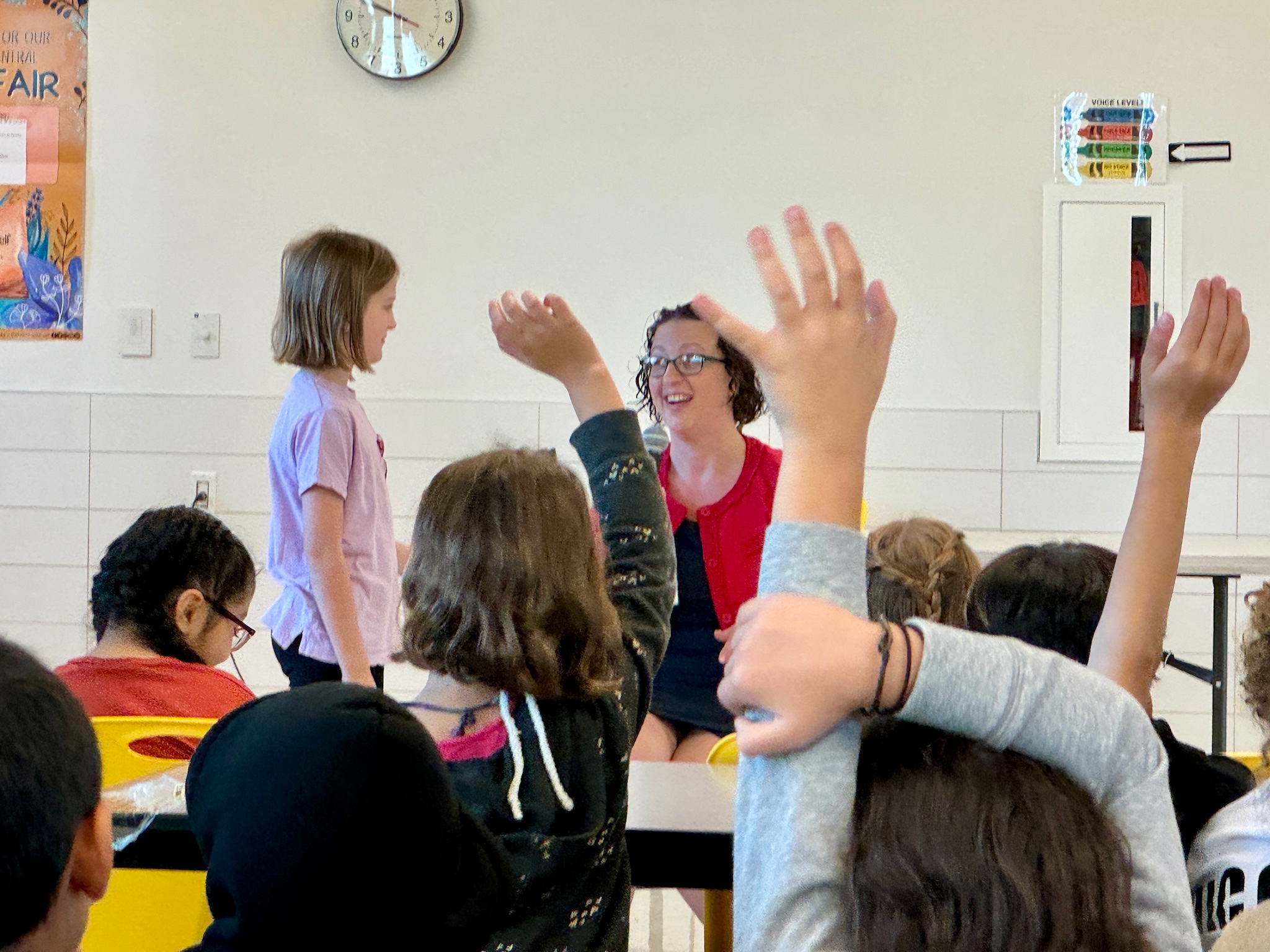 Backs of kids heads with their hands raised while seated in cafeteria seats. Councilmember Nadeau is visible smiling, speaking to a young student standing in front of her.