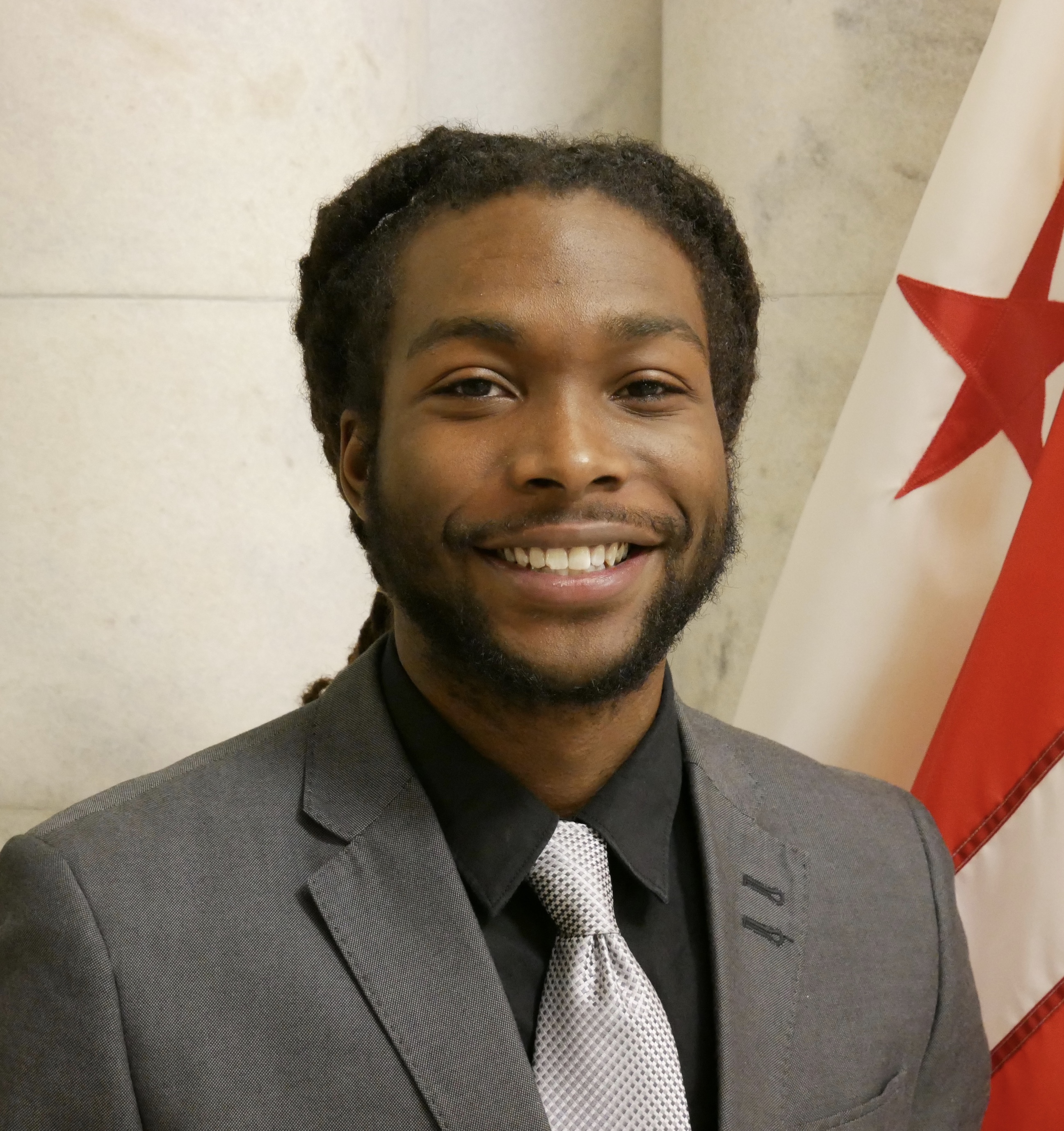DayVon Fuller smiling, stands in front of a D.C. flag, wearing a gray suit and tie.