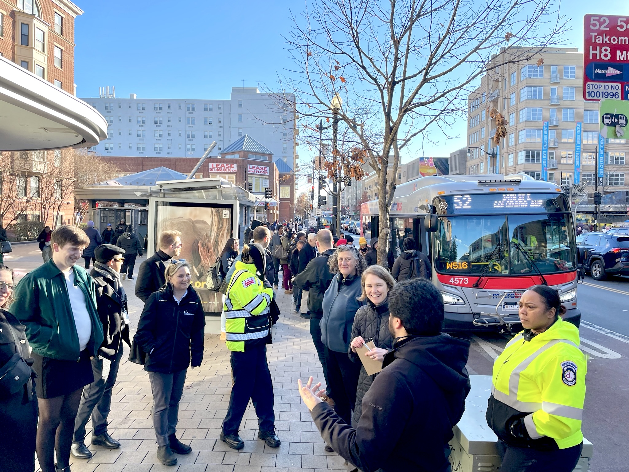 A group of residents, wearing coats on a chilly day, on the sidewalk talking. One man gestures with his hands and is speaking. Two women in yellow safety jackets/uniforms are present. A 52 bus is pulled up at the curb.