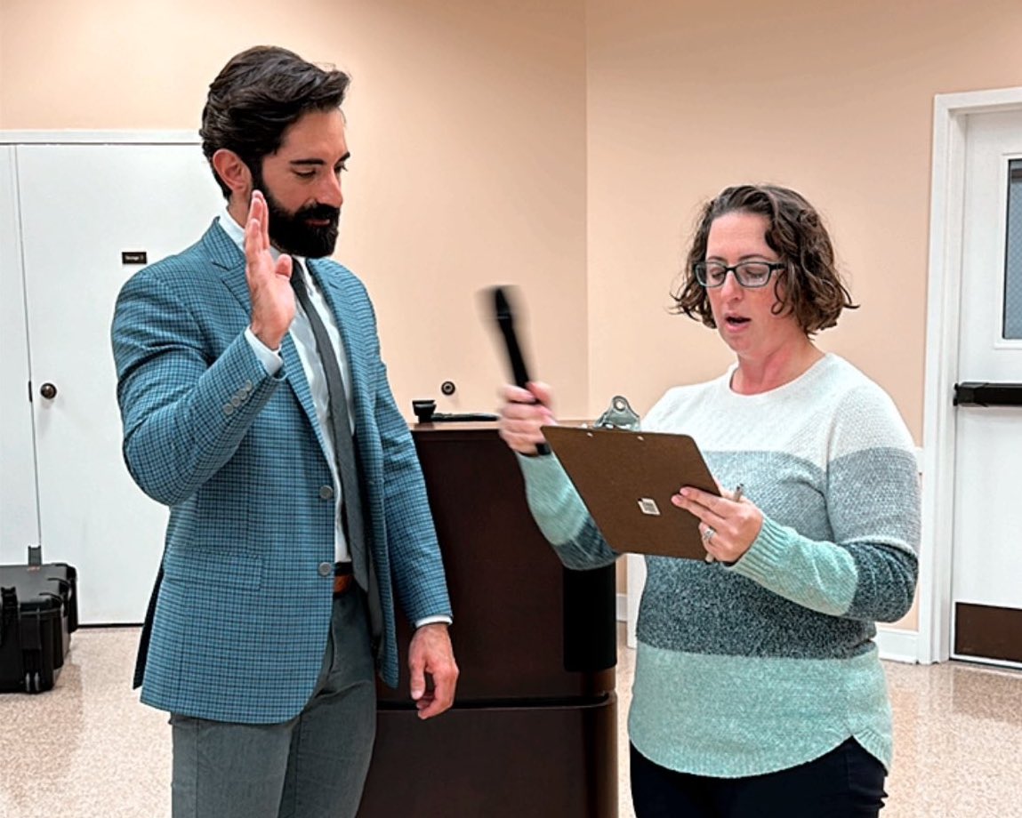New ANC 1B06 Commissioner Miguel Trindade Deramo, wearing jacket and tie, raises his right hand as Councilmember Nadeau, holding a microphone and reading off a clipboard, administers the oath of office.