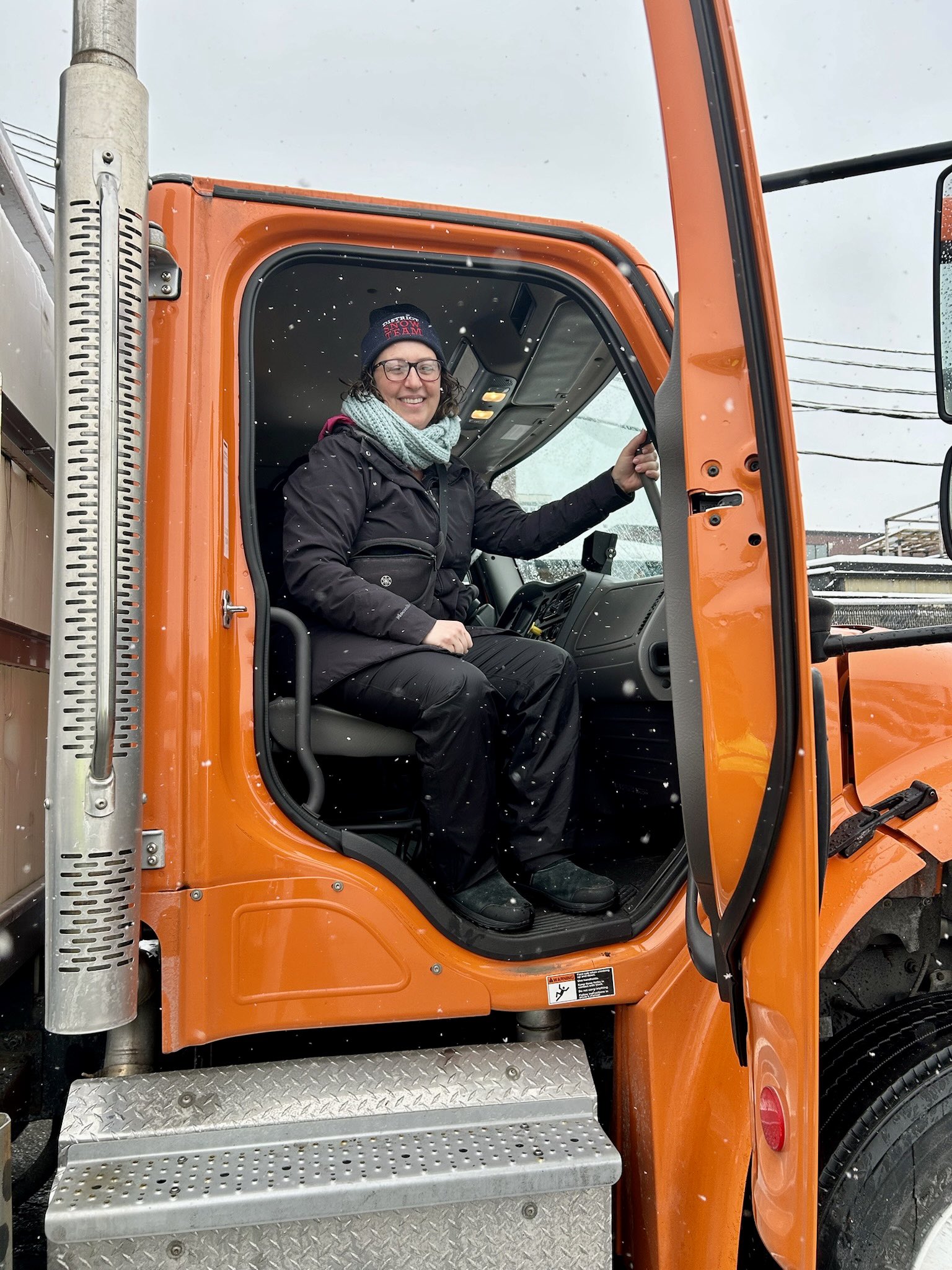 Councilmember Nadeau sits in the passenger side of a parked snow plow, with the bright orange door opened, wearing a winter hat, scarf, winter coat, smilling broadly.