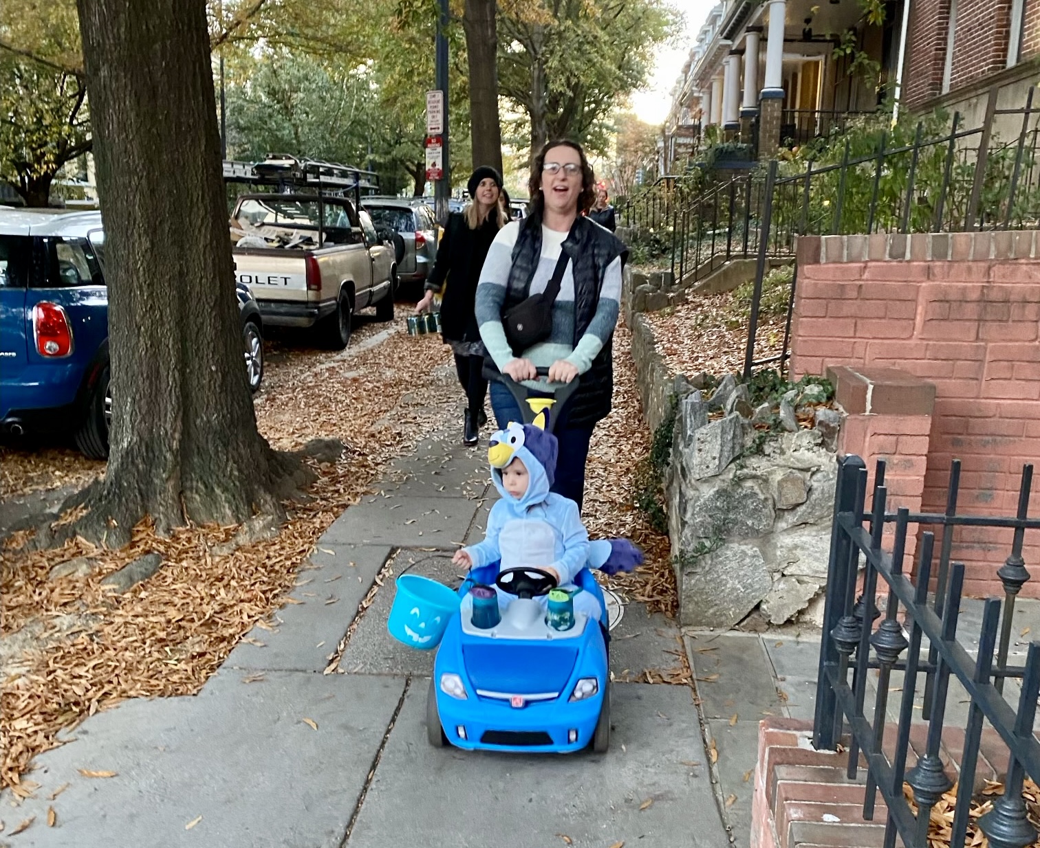 Councilmember Nadeau is pushing her daughter along the sidewalk in a toy car. Her daughter is wearing a blue bird costume and holding a blue bucket. Councilmember Nadeau is also in costume. There are leaves along the sidewalk and cars parked alongside it.